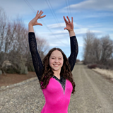 A smiling girl with long, curly brown hair strikes a dance pose with her arms raised high on a gravel path. She wears a bright pink leotard with black lace sleeves and sparkling silver trim. Bare trees and a blue sky with soft clouds fill the background.