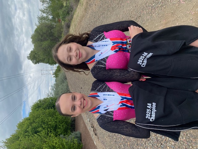 A large group of female gymnasts of various ages poses together in a gym. Most of the girls wear bright pink leotards, while a few wear black and pink designs. They are arranged in three rows against a plain white wall, with some sitting on a blue gym mat and others standing.
