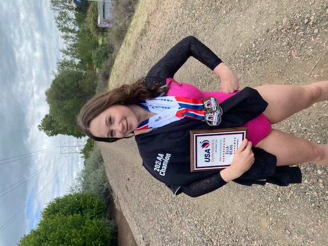 A young gymnast stands on a gravel path, smiling while wearing a pink and black leotard and multiple medals around her neck. She holds a black jacket with the white text 2025 AA Champion and a plaque from USA Gymnastics Washington that honors Ella Bean as a 2025 Graduating Senior.